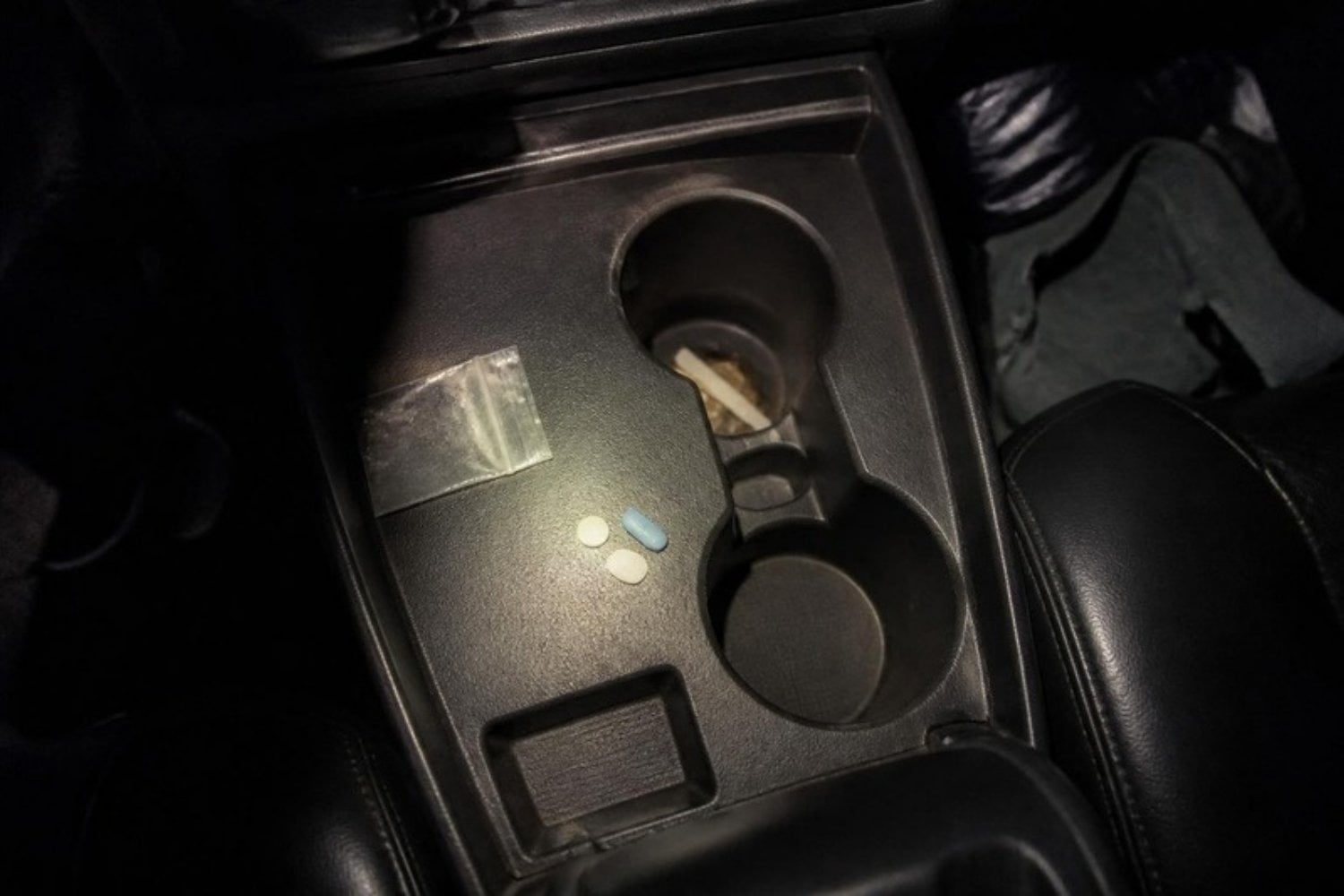 A small clear baggie and a few pills on a car's center console at night, flashlight glow