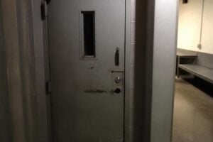 A county jail holding cell door with a small window, concrete floor, harsh light