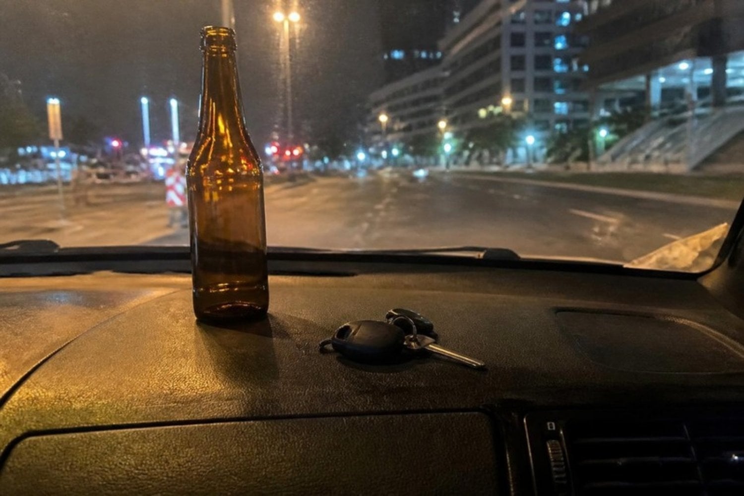 Car keys next to an empty beer bottle on a car dashboard at night, city lights through the windshield