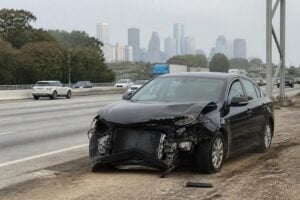 A damaged sedan pulled onto the shoulder of a Houston freeway after a crash, downtown skyline hazy in the distance