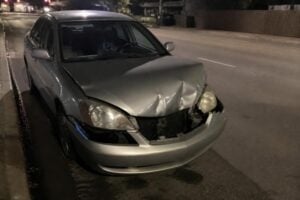 A rideshare-style sedan with a crumpled front bumper at a Houston curb at night, phone mount on the dashboard